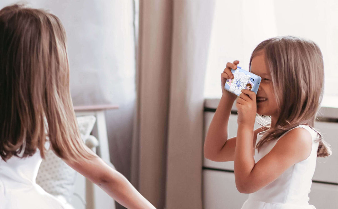 Girl using Disney Frozen digital camera for kids with 2 inch color screen and Frozen graphics, taking photos of another child in white dress.