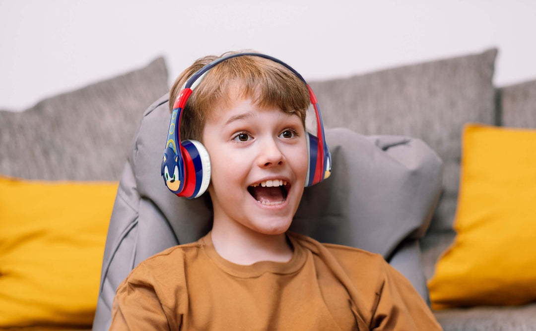 Child wearing Sonic The Hedgehog Bluetooth headphones, smiling and enjoying high quality stereo sound, seated on a gray couch with yellow pillows.